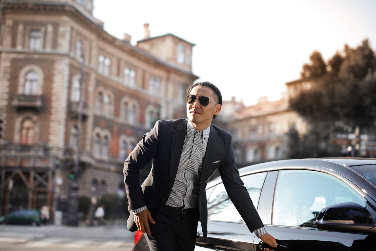 Services Confident businessman in sunglasses standing by car in city street.